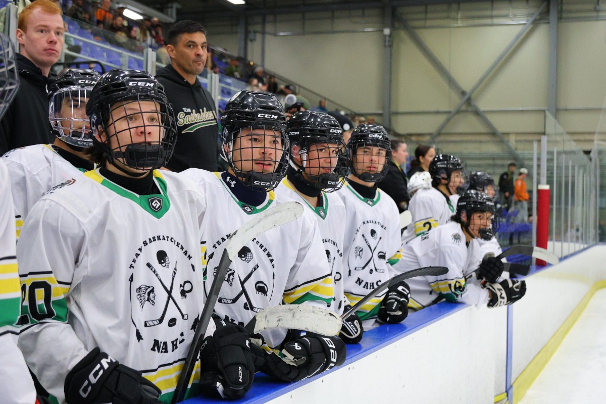 Une jeune équipe de hockey est assise sur le banc pendant un match. Les joueurs portent des maillots de la Saskatchewan. Ils regardent tous vers la patinoire, en direction de quelque chose qui se passe hors champ.